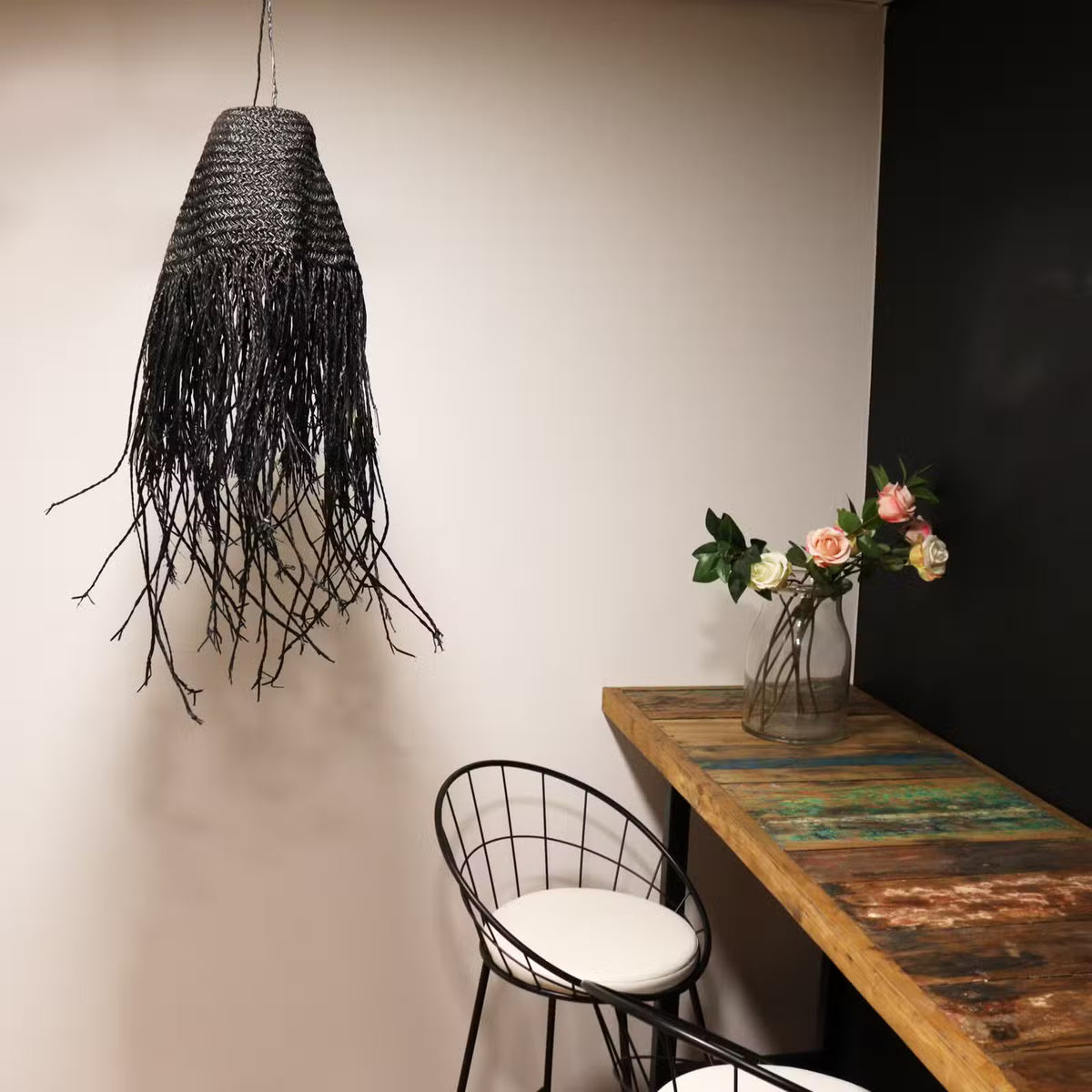 Abat-jour Sanaa en fibres noires tressées suspendu dans un intérieur bohème avec mobilier en bois et décoration végétale.