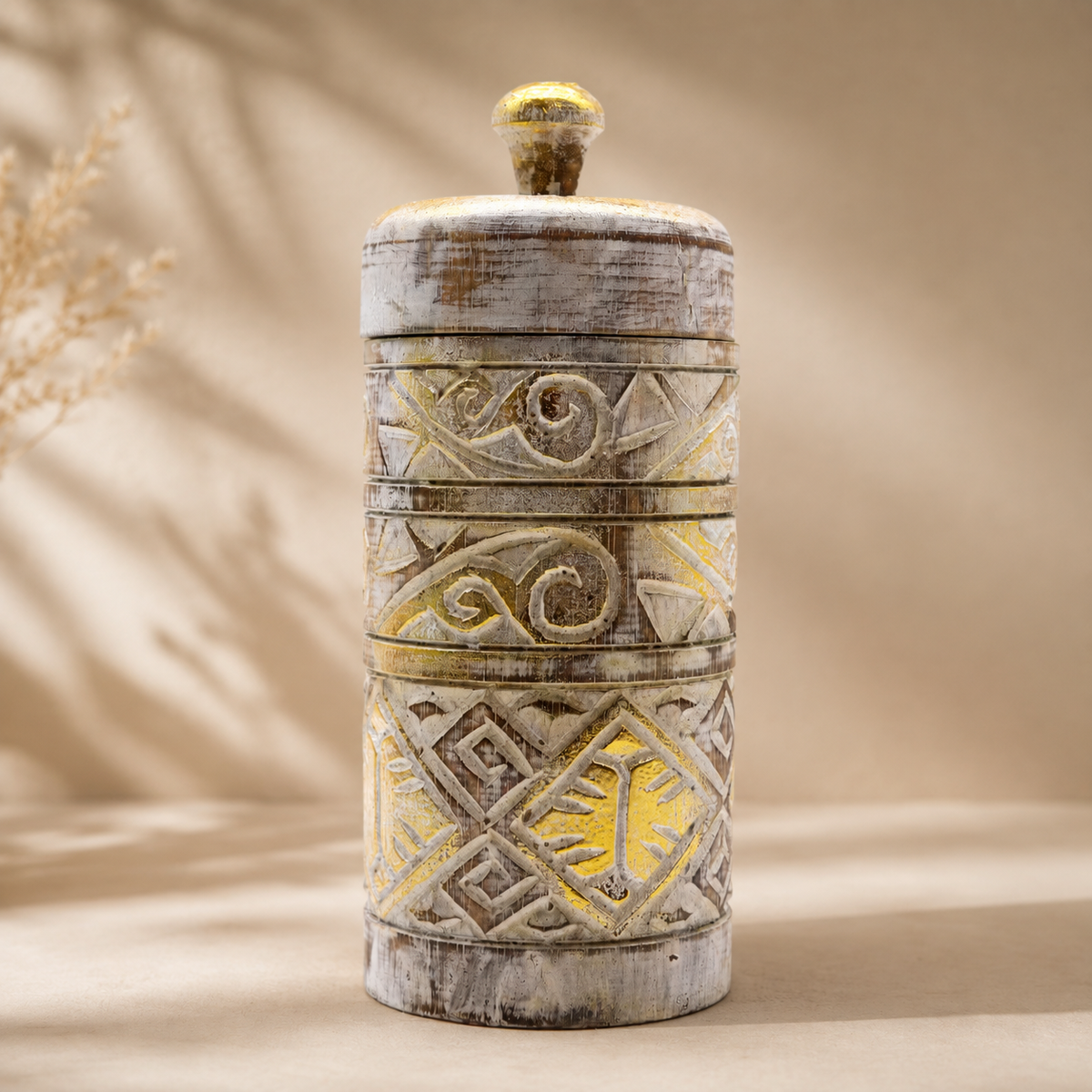 Boîte décorative Alta en blanc patiné et doré, photographiée de face sur un fond beige neutre, mettant en valeur ses motifs sculptés et sa silhouette cylindrique.