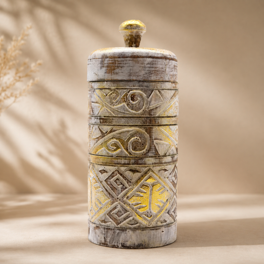 Boîte décorative Alta en blanc patiné et doré, photographiée de face sur un fond beige neutre, mettant en valeur ses motifs sculptés et sa silhouette cylindrique.