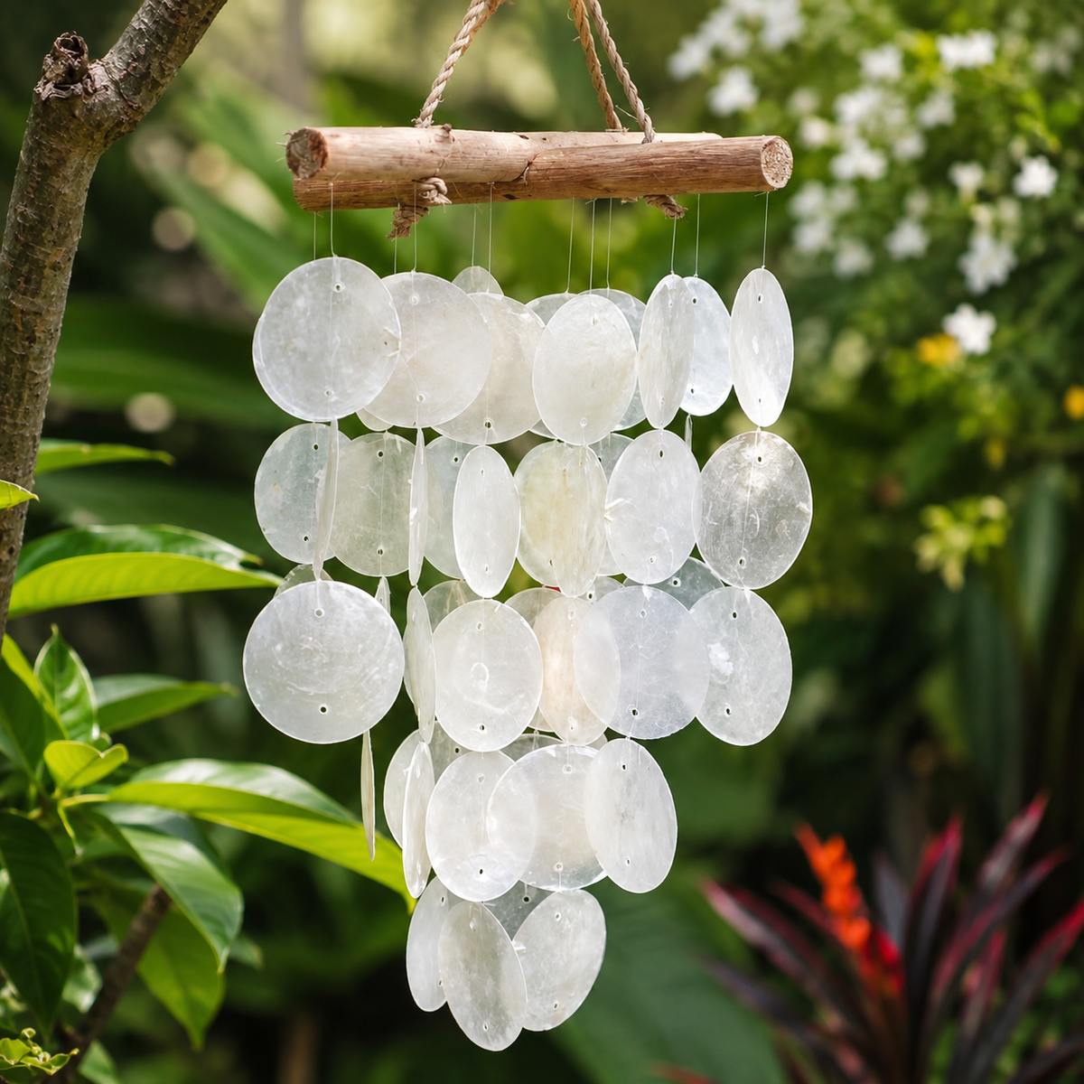 Carillon Perla en Capiz blanc et bois flotté suspendu parmi une végétation tropicale.