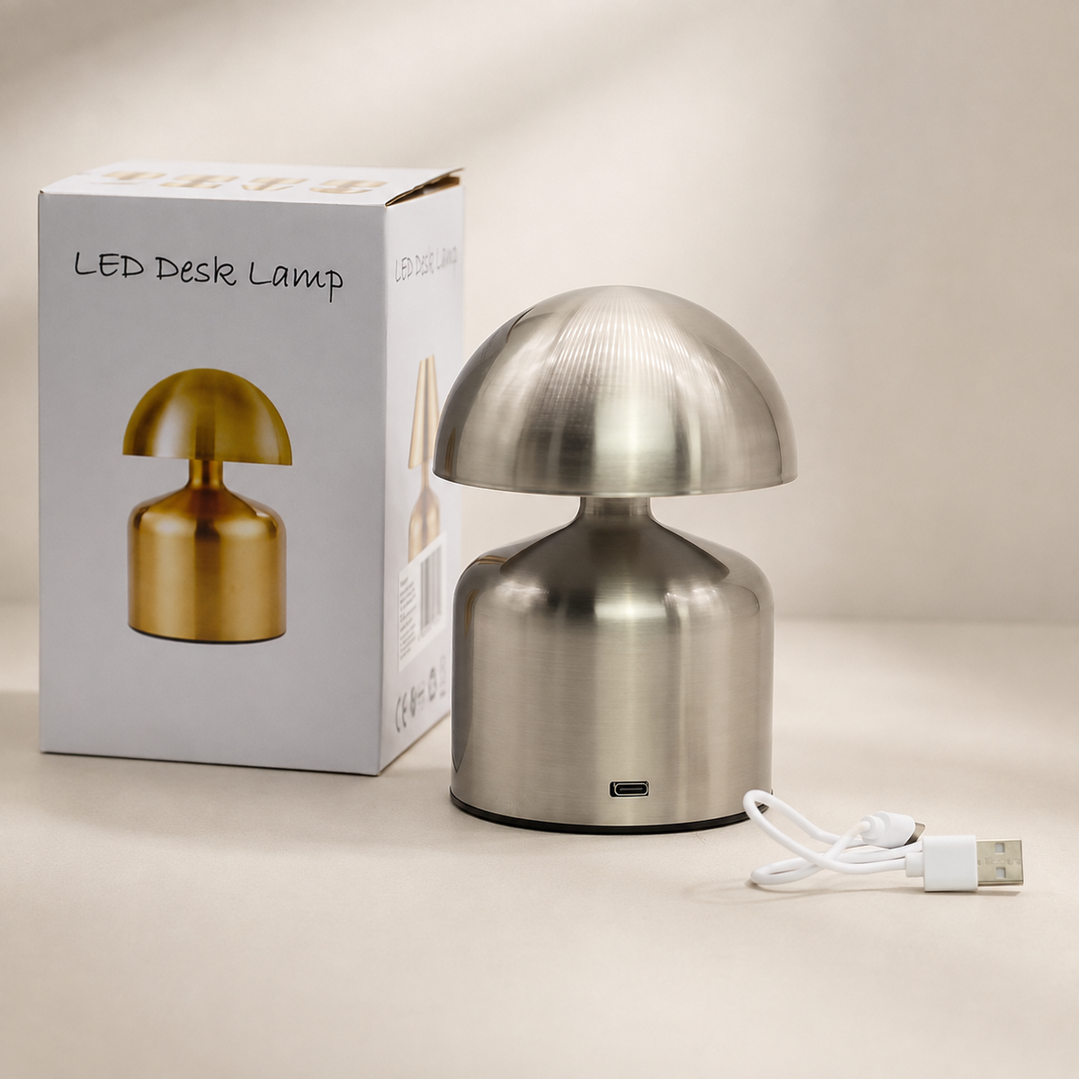 Lampe Noma argent présentée avec son emballage et son câble USB sur fond beige neutre.