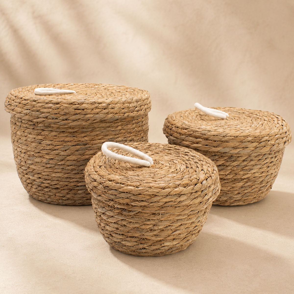 Trio de paniers Liora en fibres naturelles tressées avec couvercles et poignées blanches, présenté sur fond beige.