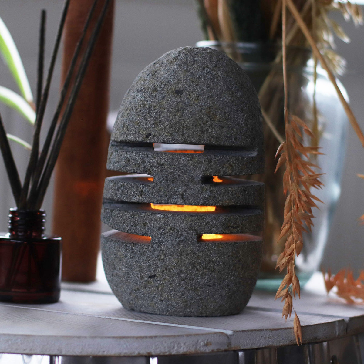 Photophore Néra en pierre de rivière allumé dans une ambiance chaleureuse, posé près d’objets décoratifs, avec une lumière tamisée qui met en valeur ses fentes lumineuses.